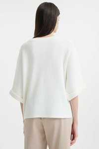 Maglione bianco texturizzato con maniche ampie, polsini arrotolati e una forma rilassata, abbinato a pantaloni beige. Il design enfatizza il comfort.