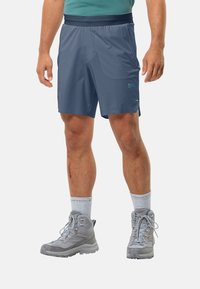 Herren Navy-Synthetik-Shorts mit elastischem Bund, seitlichen Taschen und reflektierendem Logo. Getragen mit grauen Wanderschuhen und hellgrauen Kniestrümpfen.