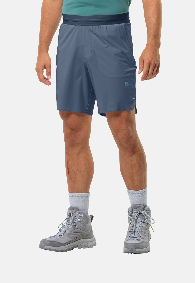 Herren Navy-Synthetik-Shorts mit elastischem Bund, seitlichen Taschen und reflektierendem Logo. Getragen mit grauen Wanderschuhen und hellgrauen Kniestrümpfen.