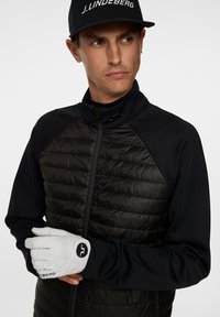 Man som bär en svart zip-jacka med en kviltad front, vit golfhandske och en svart keps med "J.LINDEBERG"-logotyp, som blickar lätt åt sidan.