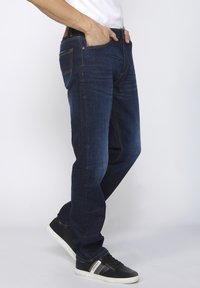 Donkerblauwe denim jeans met een rechte snit, vijf zakken, contrasterende gele stiksels en een ritssluiting, gecombineerd met zwarte sneakers.