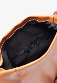 Ruskea nahkalaukku, jonka kultaketju on auki, paljastaen mustan kangasvuoren sisätaskun, jossa on teksti "ANNA FIELD."