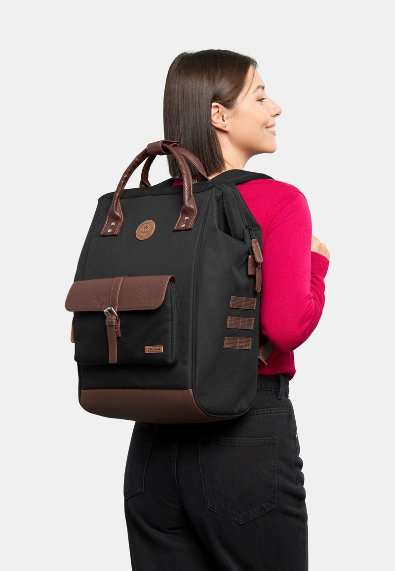 Sac à dos en tissu noir avec des accents en cuir marron, doté d'une poche avant à rabat, de deux poignées supérieures et de bretelles ajustables. Détail du logo rond visible.