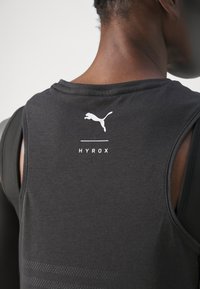 Top sportivo senza maniche nero con logo Puma bianco, testo "HYROX" e dettagli texturizzati sulla parte bassa della schiena. Tessuto morbido, vestibilità comoda.