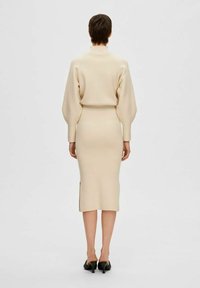 Knit dress in light beige with a high collar, puffed sleeves, and a fitted silhouette. Side slit detail and worn with black heeled shoes.