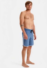 Μπλε μαγιό σορτς με υφασματικό μοτίβο, ελαστική μέση, μαύρο κορδόνι και λευκές πλαϊνές λεπτομέρειες. Το μοντέλο στέκεται ξυπόλητο και γυμνόστηθο.