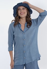 Camicia button-up di lino azzurro chiaro con maniche arrotolate, abbinata a un cappello a secchiello in crochet blu scuro. Texture liscia, vestibilità rilassata.