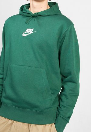 Osoba ubrana w zieloną bluzę Nike z kapturem i przednią kieszenią oraz beżowe spodnie, widoczna od szyi do talii.