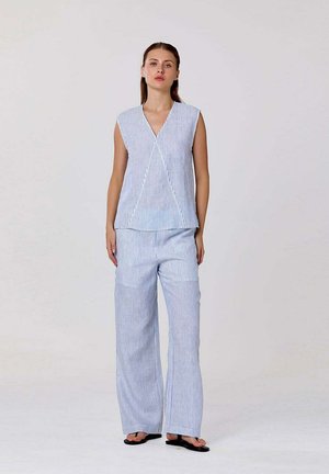 Femme debout portant un haut sans manches rayé vertical bleu et blanc et un pantalon large assorti avec des sandales noires sur fond blanc.