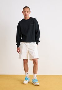 Schwarzer Sweater mit weißem Logo, weiße Shorts und Sneakers mit blauen Akzenten. Glattes Material und normale Passform, getragen mit weißen Socken.