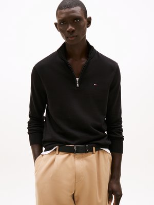 Maglione nero con collo a zip, maniche lunghe e polsini a costine, abbinato a pantaloni beige e una cintura nera con fibbia in argento.