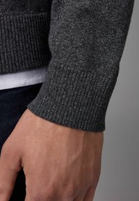Mörkgrå stickad tröja med ribbad manschett; närbild av slät textur och organisk form; bärs över ett vitt lager under.