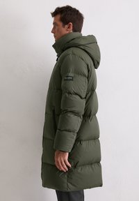 Donkergroene pufferjas met capuchon, met een doorgestikt ontwerp, ritssluitingen aan de zakken en elastische manchetten. Gemaakt van lichtgewicht, geïsoleerd materiaal.