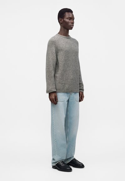 Homme debout portant un pull en tricot gris, un jean large bleu clair et des mocassins en cuir noir sur un fond blanc uni.