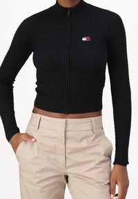Top corto de punto acanalado negro con cremallera y mangas largas, que presenta un pequeño logo de Tommy Jeans en el pecho. Combinado con pantalones beige.