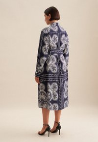 Abito blu navy con motivi paisley bianchi, maniche lunghe, colletto e una cintura in vita. Indossato con sandali neri con tacco alto.