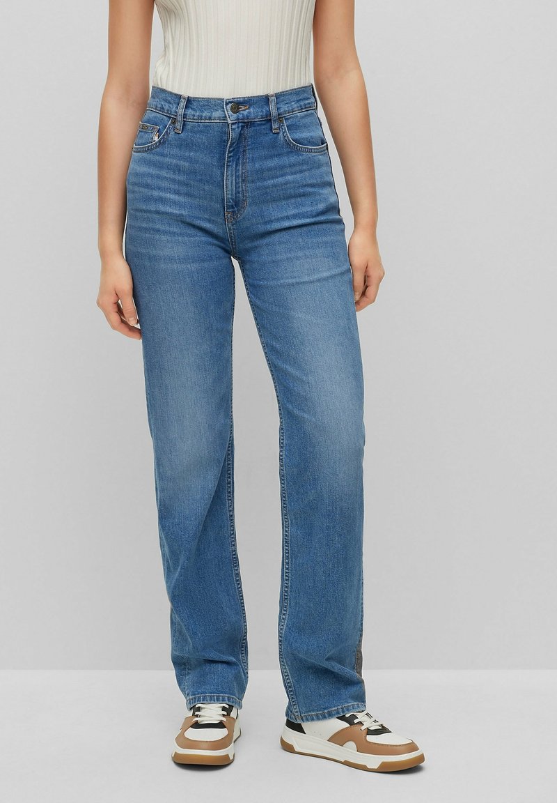 Jeans taille haute bleu clair avec une coupe droite, cinq poches et un délavage subtil. Associés à des baskets blanches et marron.