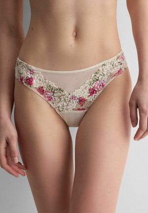 Mujer con bragas beige de encaje con bordado floral en rosa y verde, de pie con las manos relajadas a los lados.