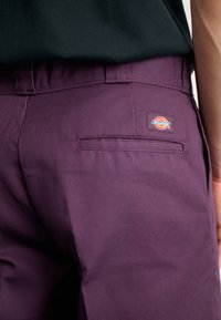 Pantaloni mov dintr-un material rezistent, cu un buzunar pe spate și un etichetă cu marcă. Au o textură netedă și o croială ajustată.