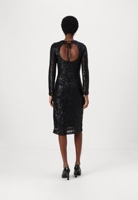 Robe noire à sequins avec des manches longues, un dos nu maintenu par un ruban, et une longueur jusqu'aux genoux. Texture lisse, silhouette ajustée.