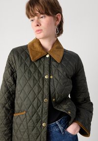 Gequilted olijfgroene jacket met een tan corduroy kraag, gouden drukknoopjes en zijzakaccenten. De stof lijkt glad met een diamantpatroon.