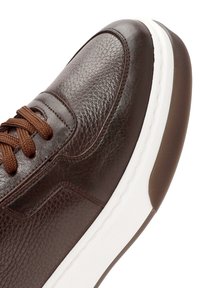 Zapatilla de cuero marrón con un acabado texturizado, que presenta detalles de cordones marrones, una entresuela blanca y una suela de goma.