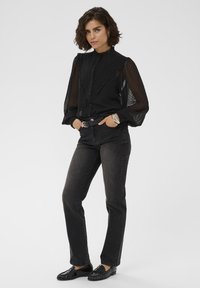 Woman with short curly hair wearing a black sheer dotted blouse, dark jeans, black belt, silver bracelets, and black loafers, standing with hands in pockets.