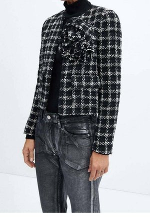 Czarna i biała krótka kurtka w kratę z dużą kwiatową aplikacją z materiału, noszona na czarnym golfie i wyblakłych czarnych dżinsach.