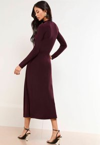 Rochie lunga burgundă cu mâneci lungi, corset ajustat, fustă fluidă și material neted; asortată cu pantofi cu toc înalt negri, cu bretele.