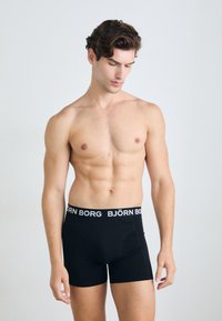 Fekete boxer alsónadrág széles elasztikus derékkal, amelyen a "BJÖRN BORG" logó található. A modell izmos testalkatú, meztelen torso-val oldalról pózol.