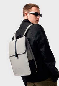 Light gray backpack with a black strap closure; sleek design, rectangular shape, smooth texture, featuring a contrasting black panel on the bottom.
