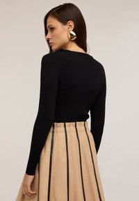 Femeie cu păr lung și brunet poartă un pulover negru cu textură în relief, o fustă bej cu dungi verticale negre și cercei mari din aur, îndreptată spre stânga.