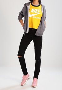 Osoba oblečená v žlutém sportovním tričku Nike, šedé zipové mikině, roztrhaných černých džínách a růžových teniskách Nike, stojí s rukama v kapsách.