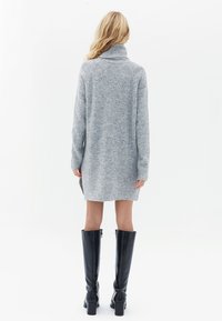 Abito in maglia grigia con colletto alto, texture a costine e silhouette dritta, abbinato a stivali neri lucidi fino al ginocchio.