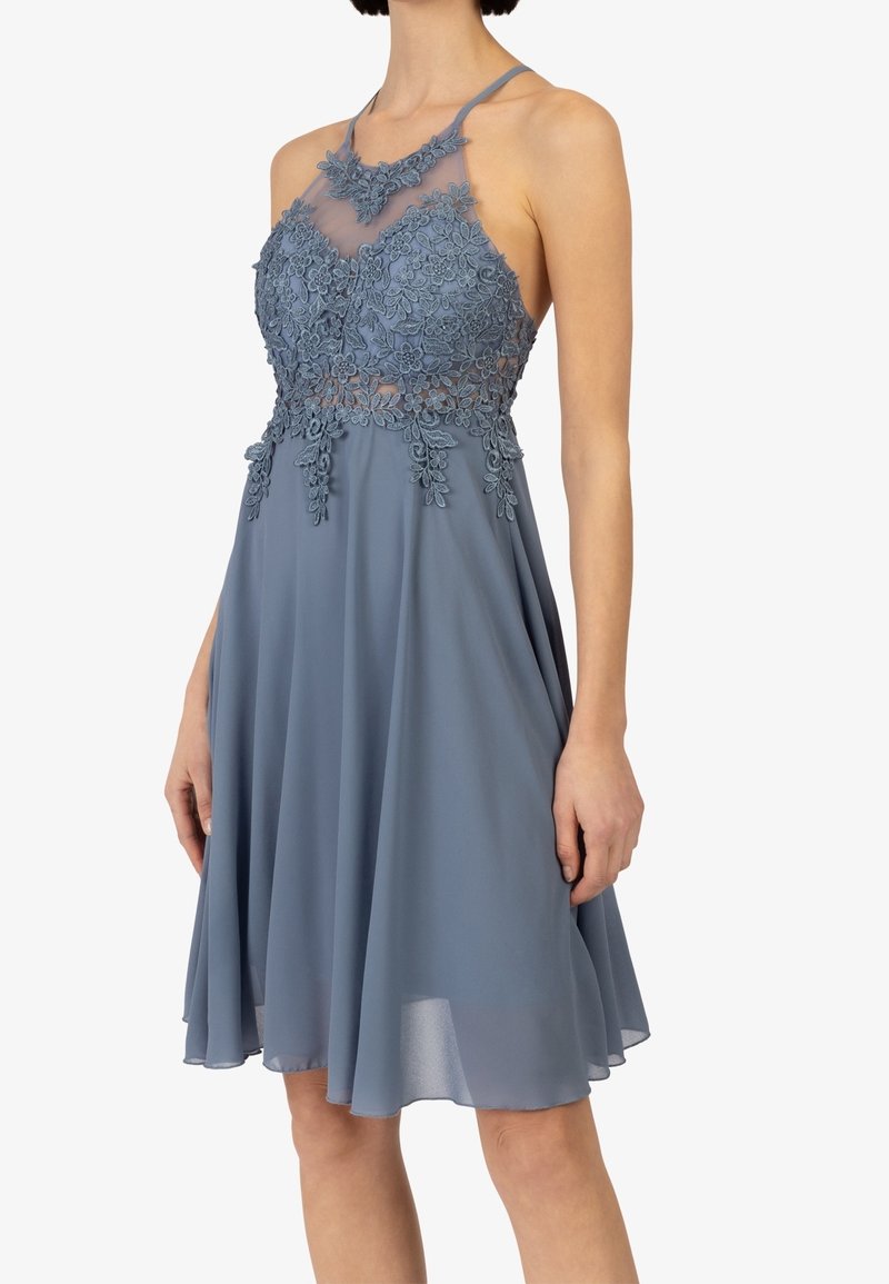 Robe bleue sans manches longueur genou avec corsage en dentelle florale, encolure transparente et jupe fluide portée par une femme debout.
