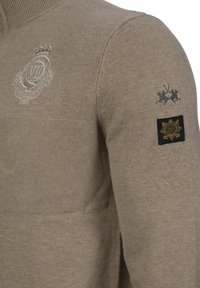 Beżowy robiony na drutach sweter z wysokim kołnierzem, z teksturowanymi wzorami, wyhaftowanym herbem oraz czarną łatą z złotymi detalami na rękawie.