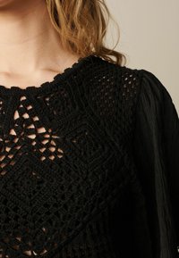 Top in pizzo nero con un design a motivo intricato e maniche a sbuffo texturizzate. Le aperture a forma di chiave creano un effetto a strati.