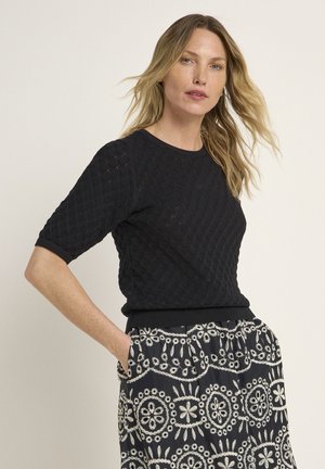 Schwarzer gestrickter Kurzarmpullover mit Rautenmuster, der über einen runden Ausschnitt und einen gerippten Saum verfügt. Kombiniert mit einem gemusterten Rock in Schwarz und Weiß.