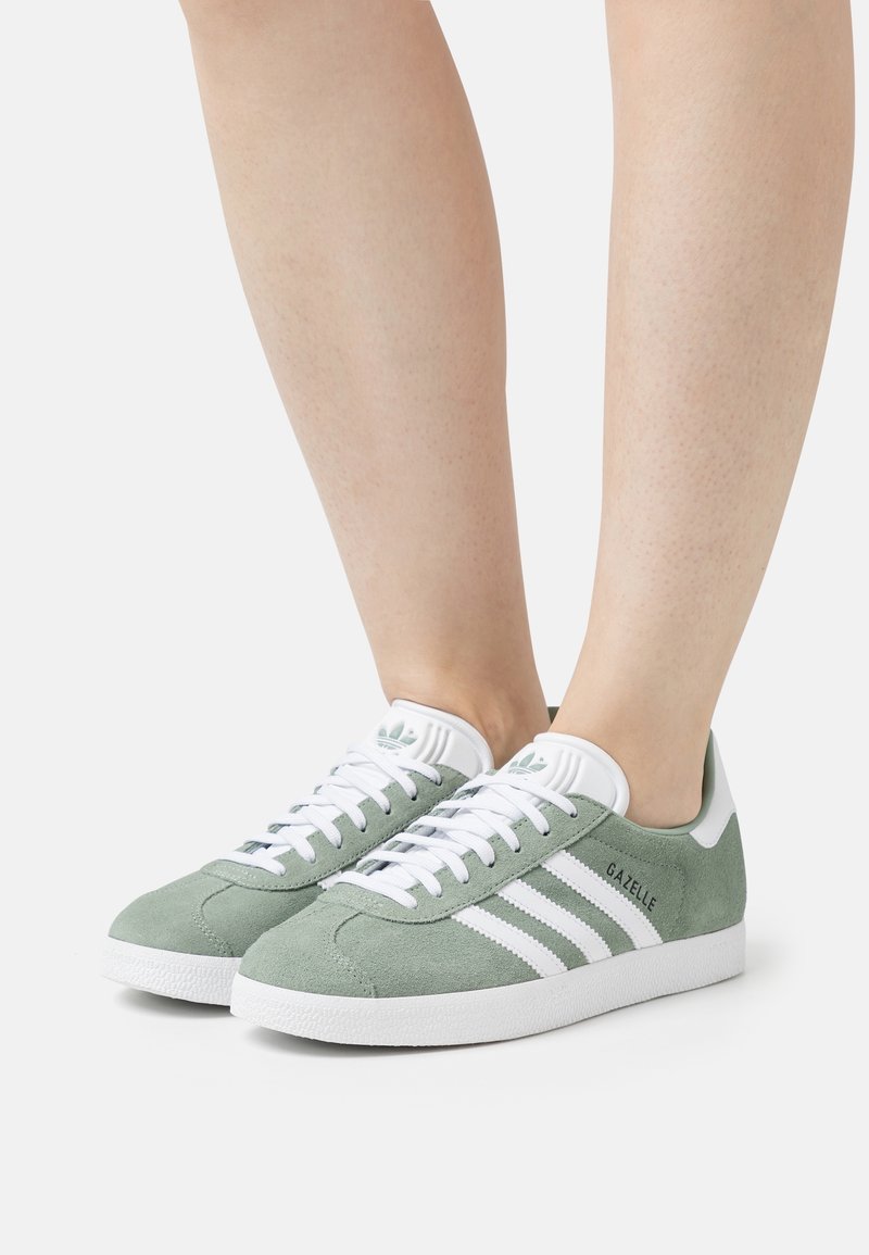 Zelené semišové tenisky s bílými akcenty, obsahující tři pruhované detaily, texturovanou podrážku a branding blízko paty. Gumová podrážka.