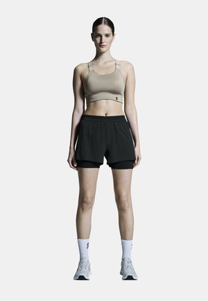 Beige Sport-BH mit verstellbaren Trägern, kombiniert mit schwarzen Shorts mit Innenslip. Graue Sportschuhe und weiße Socken.