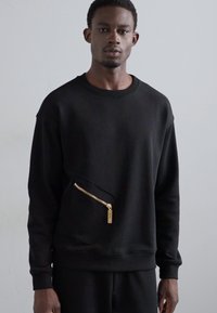 Čierna mikina vyrobená z textúrovanej látky, má predné vrecko na zips so zlatým kovovým doplnkom a okrúhly výstrih.