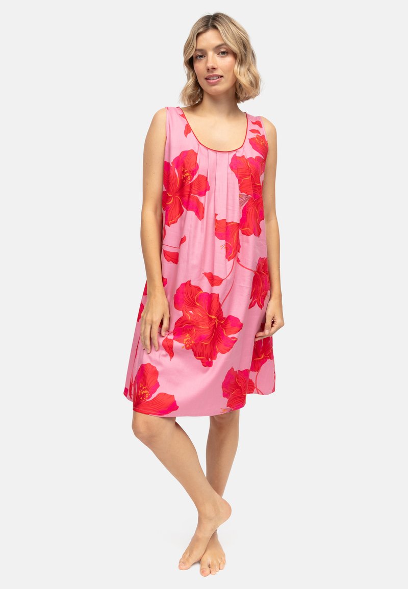 Frau steht barfuß und trägt ein ärmelloses rosa Kleid mit großen roten Blumenmustern vor einem einfarbigen hellen Hintergrund.