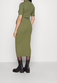 Kobieta ubrana w oliwkową, dopasowaną sukienkę midi o prążkowanej fakturze z krótkimi rękawami oraz czarne, masywne botki na obcasie, stojąca przy gładkiej ścianie.