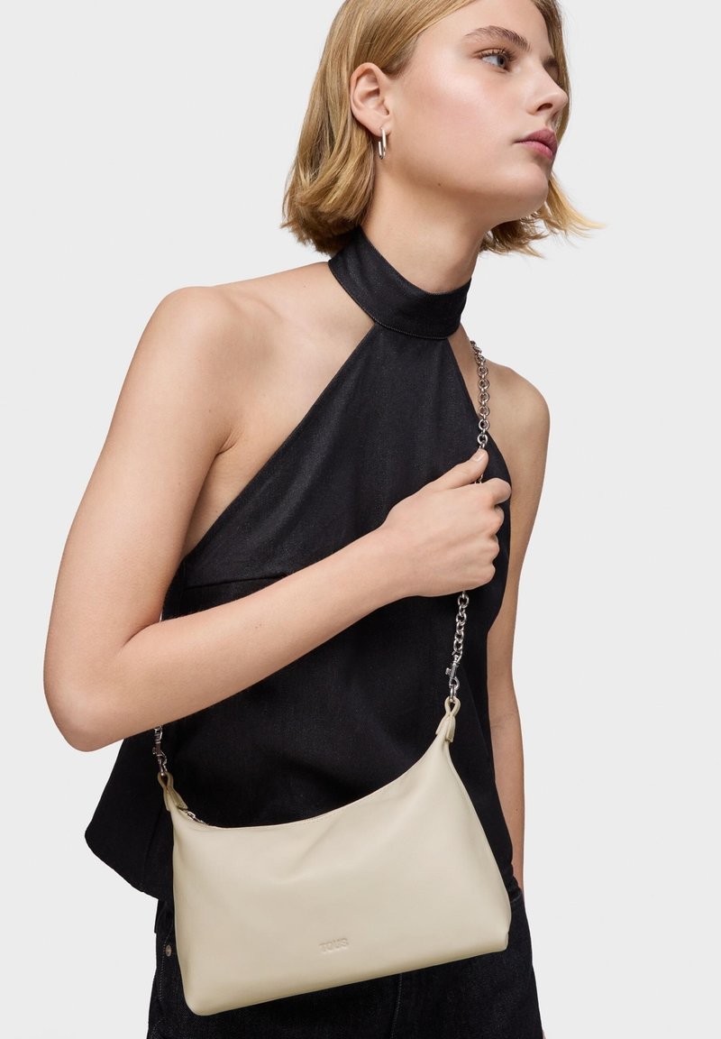 Joven mujer con top negro de tipo halter sosteniendo un bolso de cuero beige con cadena plateada sobre el hombro, contra un fondo liso.