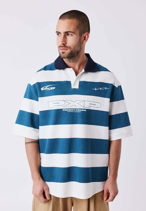 Uomo con capelli corti e barba indossa una polo larga a righe blu e bianche con logo “PXP”, guarda di lato, sfondo neutro.
