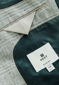 Slim fit żakiet wykonany z teksturowanej jasnoszarej tkaniny, z ciemnozielonym podszewką o subtelnym wzorze oraz metką w kolorze białym z logo.