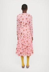 Personne aux cheveux courts portant une robe midi rose à fleurs à manches longues, des bas jaunes et des chaussures plates rayées, debout de dos.
