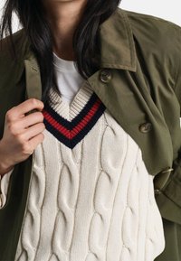 Žena upravující olivově zelený kabát, oblečená v bílém svetr vestě s copánkovým vzorem, přes modro-červený pruhovaný svetr s výstřihem do V a bílou košili.