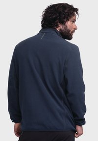 Navy fleece jacket with a high collar, featuring a subtle green accent stitch at the back and elasticated cuffs, offering a relaxed fit.