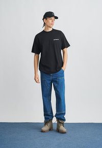 Camiseta negra de algodón con logo blanco, vaqueros azules y botas beige con cordones. El modelo posa frente a un fondo neutro.